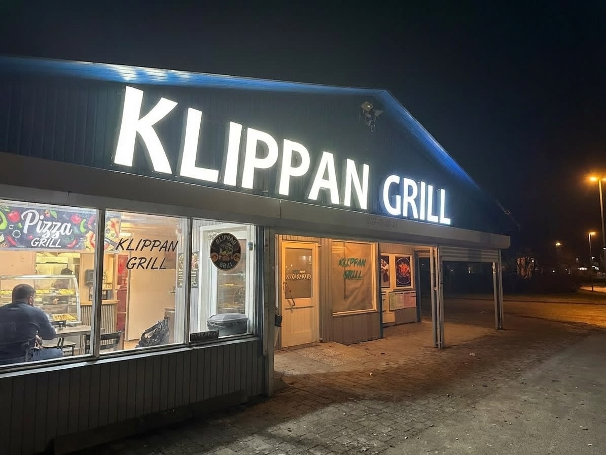 En bild tagen utomhus nattetid föreställande en byggnad med skylten "KLIPPAN GRILL" upplyst på taket. En person sitter vid ett bord inne i byggnaden, synlig genom ett fönster. Utanför byggnaden syns asfalt och gatlyktor i bakgrunden.