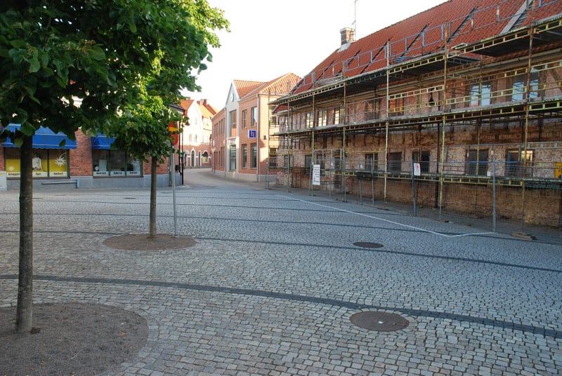 En gata belagd med gatsten i en stadsmiljö. Till vänster syns träd, butiksfasader med blåa markiser och fönster med text. Till höger står en tegelbyggnad med byggnadsställningar och ett staket. Längre bort på gatan syns fler byggnader med röda tak.