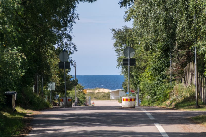 En väg skymtar fram genom en allé av gröna träd, med havet och sanddyner i bakgrunden. Två betongfundament med röda och gula markeringar blockerar vägen. Ljusblå himmel syns ovanför havet, och på vänster sida finns en soptunna. En liten vit byggnad s