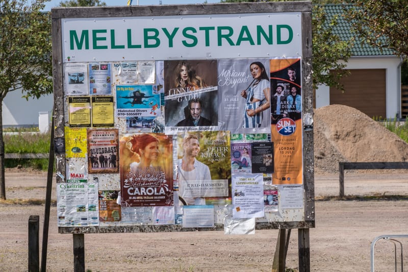 En stor, sliten anslagstavla med texten 'MELLBYSTRAND' överst, fylld med olika affischer och mindre meddelanden som annonserar evenemang och lokala tjänster. I bakgrunden syns delar av en byggnad med brunt garage och en hög med grus.
