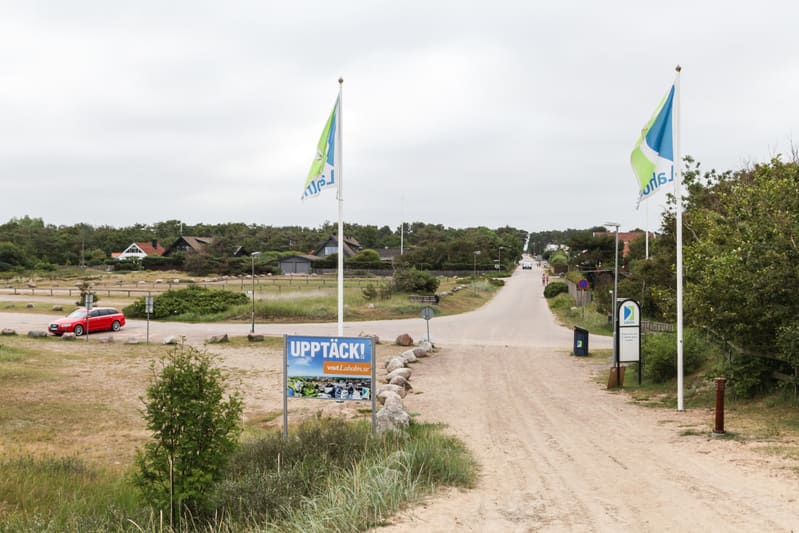 En utomhusbild som visar en sandig grusväg som mynnar ut i en asfalterad väg. Längs kanterna av vägarna finns gräs, buskar och träd. Till vänster i bilden står en röd bil parkerad. Flera hus syns i bakgrunden. Två flaggor är placerade vid sidorna av