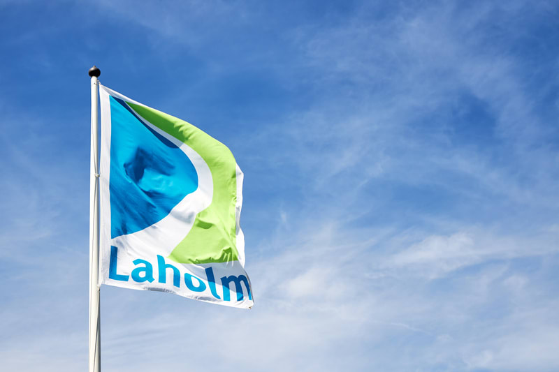 En flagga med texten "Laholm" svajar i vinden mot en klarblå himmel med tunna, vita moln. Flaggan har ett mönster i blått, grönt och vitt.