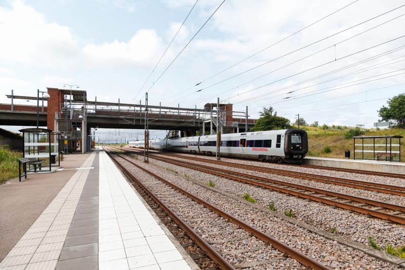 En modern station med en Oresundståg regionaltåg på spåren under en viadukt. Två plattformar med transparenta väderskydd, järnvägsräls, och grönskande buskar under en något molnig himmel.