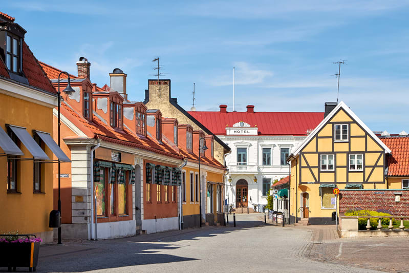 En gata med färgglada byggnader under en klarblå himmel. Till vänster syns flera hus med tegeltak och markiser framför fönstren. Längre bort på gatan finns en vit byggnad med texten "HOTEL" och ett rött tak. Till höger står ett korsvirkeshus med gul