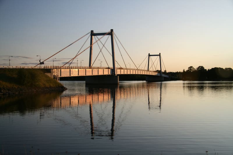 En bro med två bropelare och synliga stag som spänner över vatten. Solen lyser på brons ena sida och skapar en reflektion i vattnet. Till vänster syns gräsbevuxen mark och till höger skymtar träd vid horisonten.