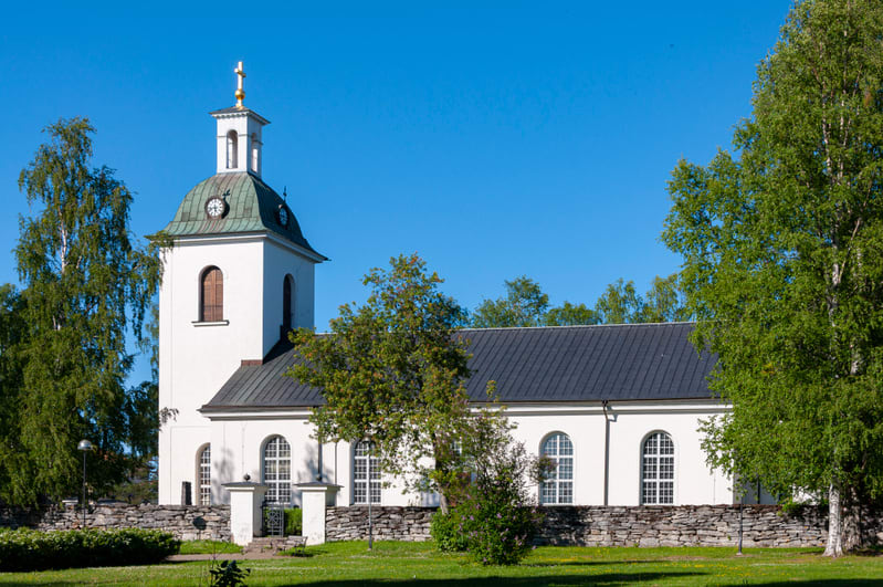 En vit kyrka med ett gråsvart tak och ett klocktorn med ett grönt tak syns under en klarblå himmel. Runt kyrkan finns gräsmattor, träd med gröna löv och en stenmur som avgränsar området framför kyrkan.