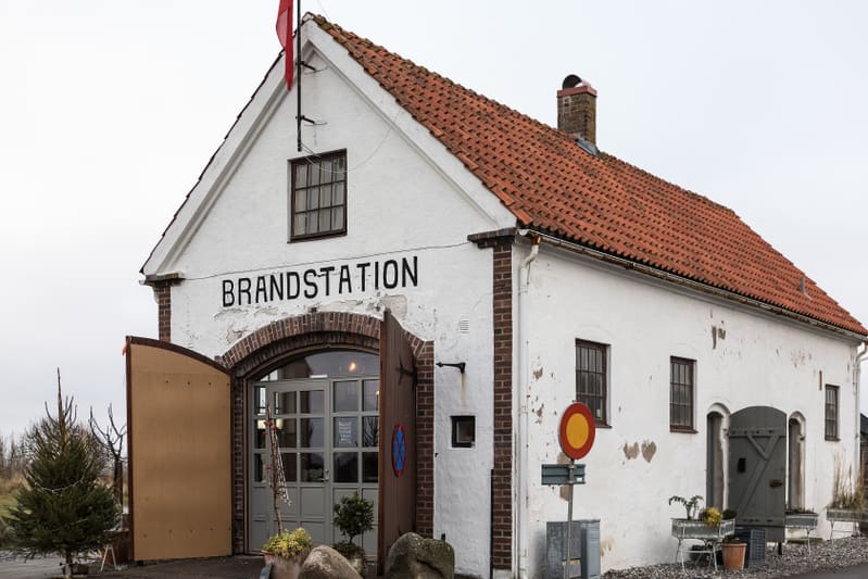 En vitputsad byggnad med rött tegeltak. På fasaden står det "BRANDSTATION" skrivet med stora svarta bokstäver. Byggnaden har en stor öppning med en brun träport samt fönster med spröjs. En julgran med belysning syns till vänster om byggnaden.