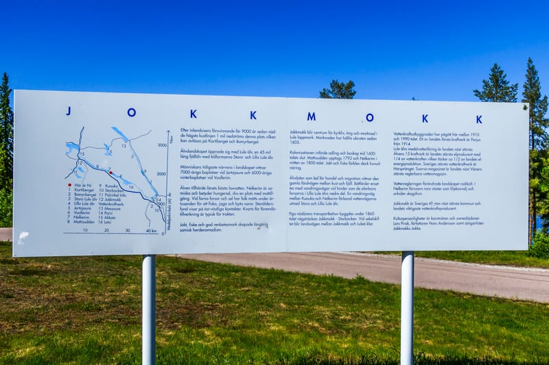 En stor, vit informationstavla med text och kartor står utomhus framför gräsfält och träd under en blå himmel. På tavlan syns texten ”JOKKMOKK” samt information om områdets historia, geografi och kultur på svenska.