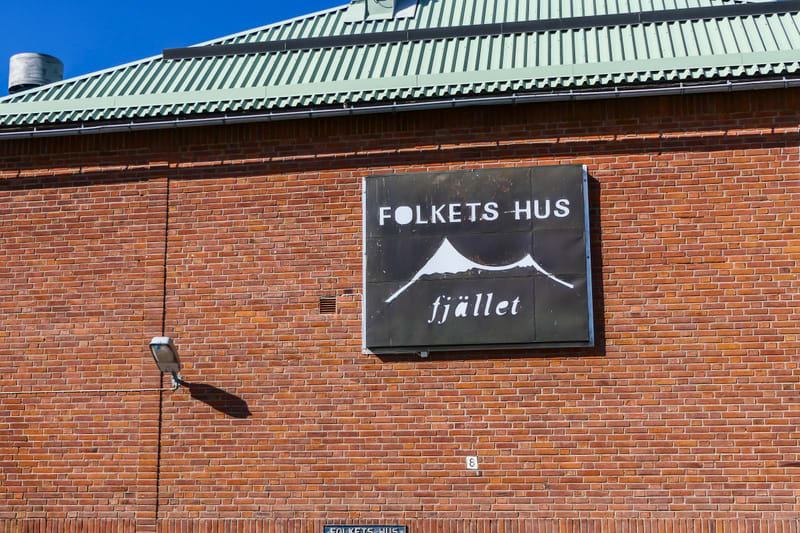 En bild av en tegelvägg på en byggnad med ett svart skylt med texten "FOLKETS HUS fjället" och en stiliserad bergstopp. Ovanför syns en del av ett grönt plåttak och en blå himmel. På väggen finns även en lampa och en liten vit nummerplatta med siffra