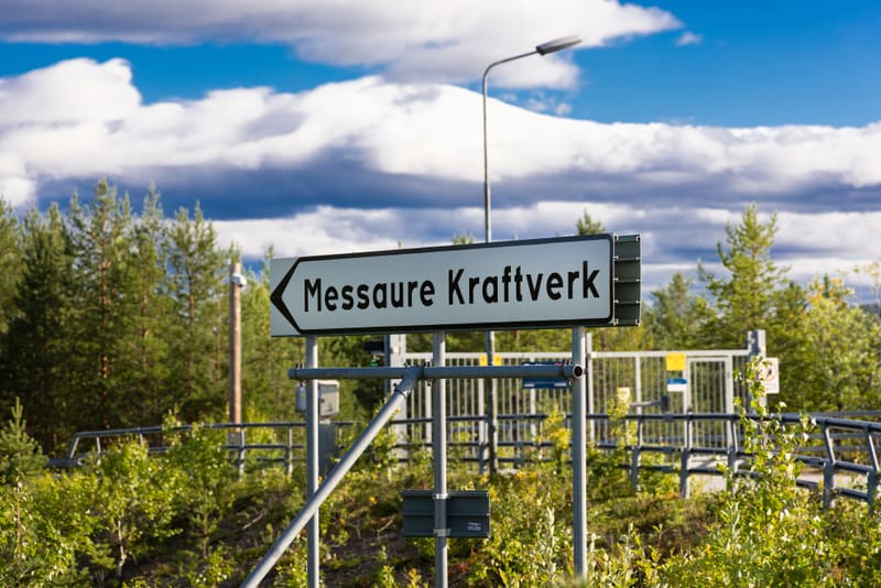 En vägskylt med texten "Messaure Kraftverk" med en pil som pekar åt vänster, står i förgrunden. Bakom skylten finns en metallgrind och en skog av barrträd under en delvis molnig himmel.