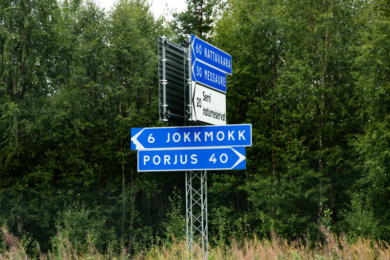 En bild som visar en vägskylt med flera blå och en vit skylt. Skyltarna anger riktningar och avstånd till orter som Jokkmokk, Porjus, Nattavaara, Messaure samt Serri naturreservat. I bakgrunden syns täta gröna träd och i förgrunden finns gräsväxter.