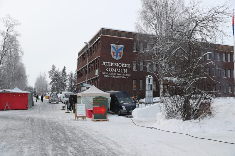 En vinterbild föreställande Jokkmokks kommunhus med snötäckta träd och mark. Utanför byggnaden står tält och fordon, och flera personer syns i bakgrunden. En skylt för "Jokkmokks kommun" och kommunvapnet är synliga på fasaden.
