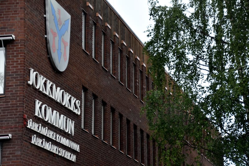 Bilden visar fasaden på en mörk tegelbyggnad med många smala fönster. En skylt med texten ”JOKKMOKKS KOMMUN” och ”JÅHKÅMÅKE KOMMUVNNA” samt ”JÅHKÅMÅKE SUOHKAN” är monterad på fasaden. Ovanför texten finns en vapensköld. Till höger i bilden syns grena