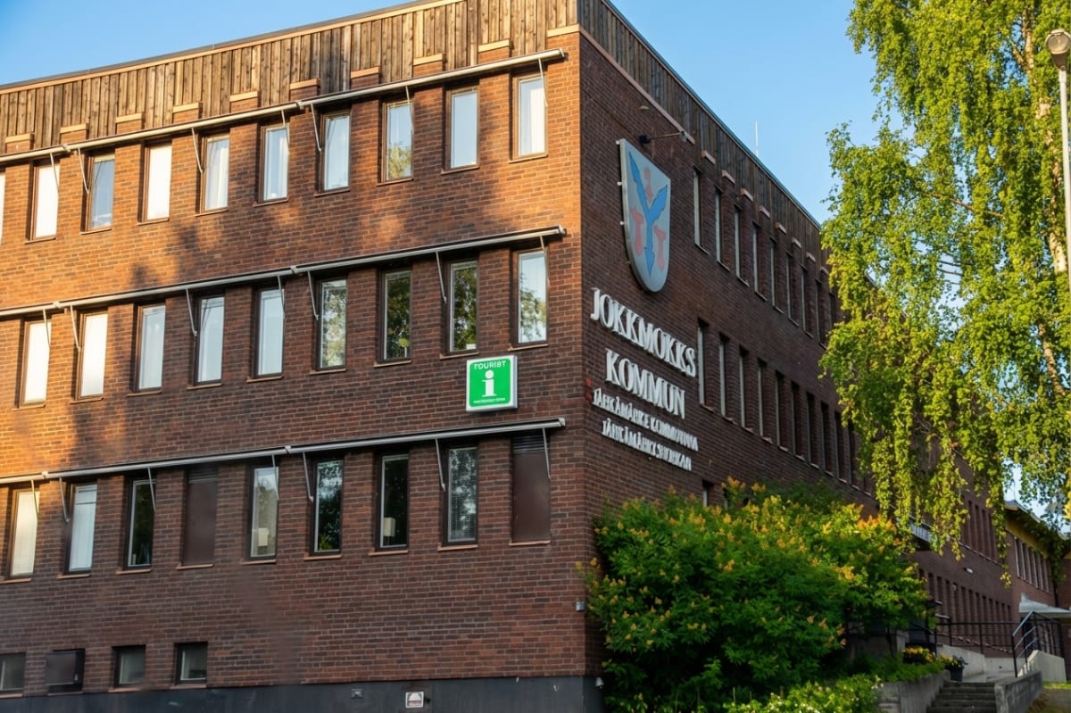 En stor, mörkbrun tegelbyggnad med många fönster och en övre fasad av trä. På byggnaden finns en skylt med texten "JOKKMOKKS KOMMUN" och dess samiska översättningar. En grön "Tourist information"-skylt syns också, samt träd med gröna löv och buskar f