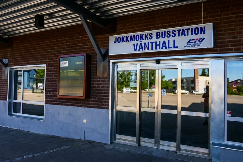 En fasad av rött tegel och ljusgrå puts med vita fönsterkarmar mot en blå himmel. Fasaden har en skylt med texten 'JOKKMOKKS BUSSTATION', en informationsskärm som visar avgångstider och en blank dörr i stål och glas. Vissa träd och en byggnad reflekt