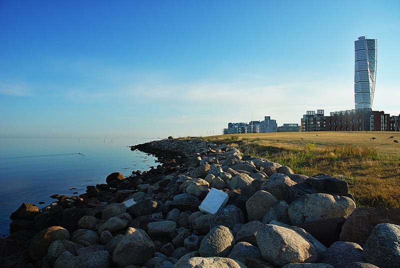 En bild som visar en kustlinje med en stenig strandkant, lugnt vatten till vänster, och en grässluttning med byggnader i bakgrunden till höger. Klarblå himmel syns ovanför.