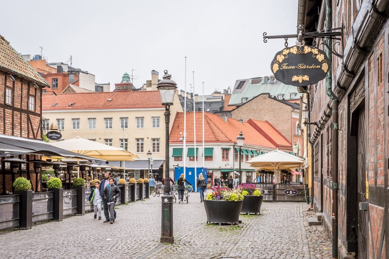 En bild av en kullerstensbelagd gata eller torg med historicistiska byggnader och uteserveringar. Flera personer promenerar och cyklar, och en skylt med texten "Faxø-Gården" är synlig. Byggnaderna har röda tak och olika fasadfärger. Planters och gatl