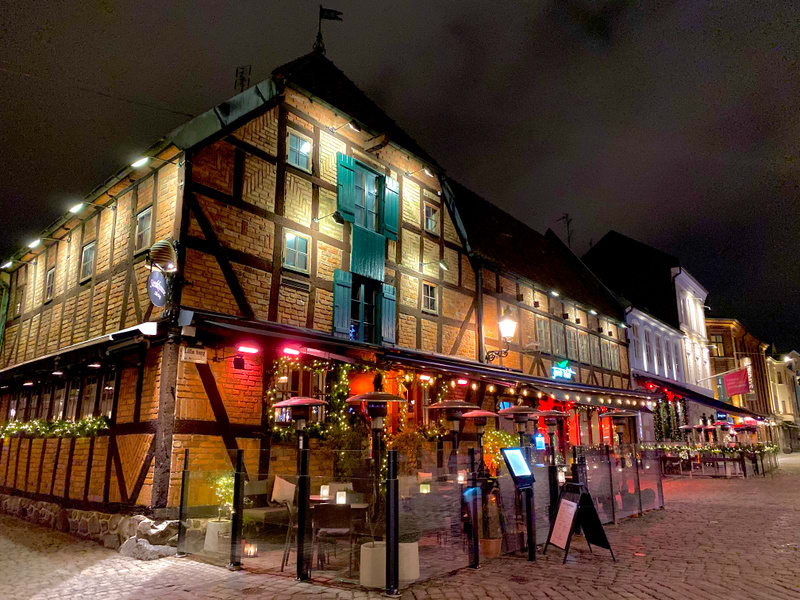 En gatsyn från Gamla Väster i Malmö under kvällstid, med en tegelbyggnad med korsvirke i fokus. Utanför finns en uteservering med infravärmare och julbelysning, samt flera skyltar och ett kullerstenslags torg. Hela scenen är upplyst i mörkret.
