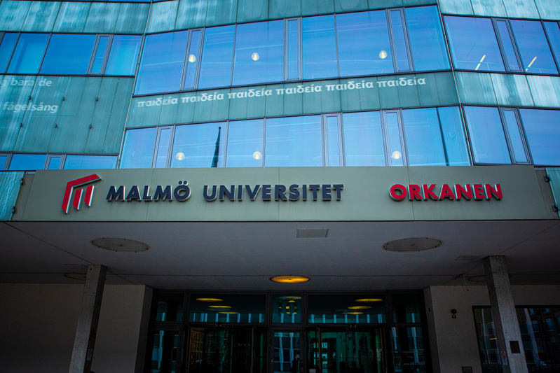 En bild som visar skylten "MALMÖ UNIVERSITET ORKANEN" över en ingång till en modern byggnad med glasfasad. Reflektioner från lampor är synliga i fönstren.