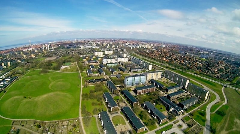 En vidvinkelbild från luften visar ett grönt parkområde med mindre kullar i förgrunden, omgivet av flerbostadshus av varierande storlek och bostadsområden med småhus. I horisonten syns en större stadskärna intill ett vattenområde under en ljusblå him