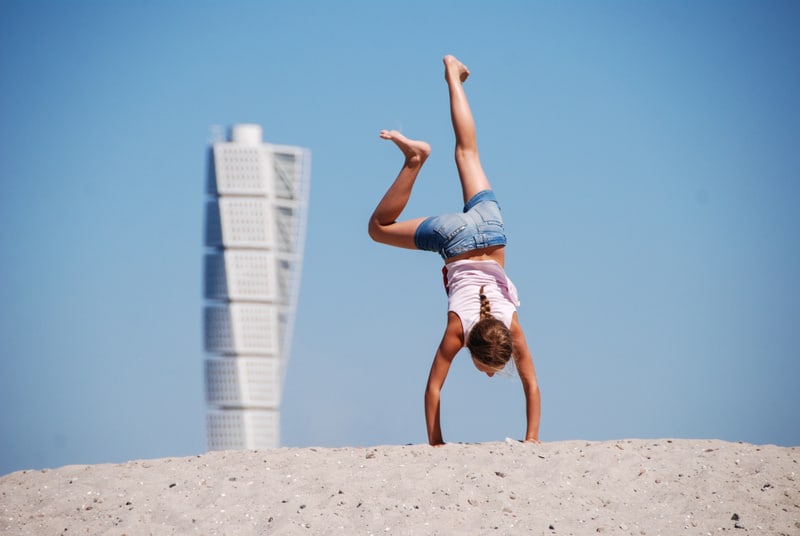 En flicka i en rosa t-shirt och blå denimshorts gör en handstående pose på en sandig yta. Hon har en fläta i håret. I bakgrunden syns en vit, modern byggnad under en klarblå himmel. Bilden är tagen utomhus i starkt solljus.