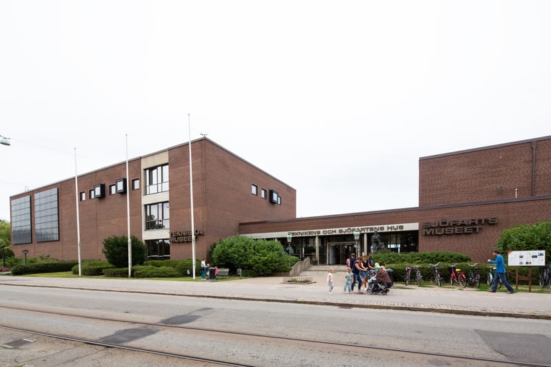 En stor, tegelröd byggnad med texten "TEKNISKA MUSEET" och "SJÖFARTS MUSEET" på fasaden, sett från gatan. Utanför byggnaden syns buskar, lyktstolpar, cyklar och några personer. På gatan syns spårvagnsspår.