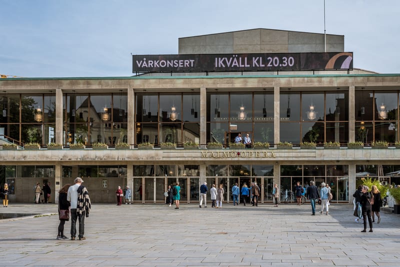 En fasadbild av Malmö Opera med en skylt som visar "VÅRKONSERT IK KVÄLL KL 20.30". Flera människor rör sig på torget framför byggnaden och vissa går in genom entréerna. Växter syns i fönsterkarmar och i förgrunden.