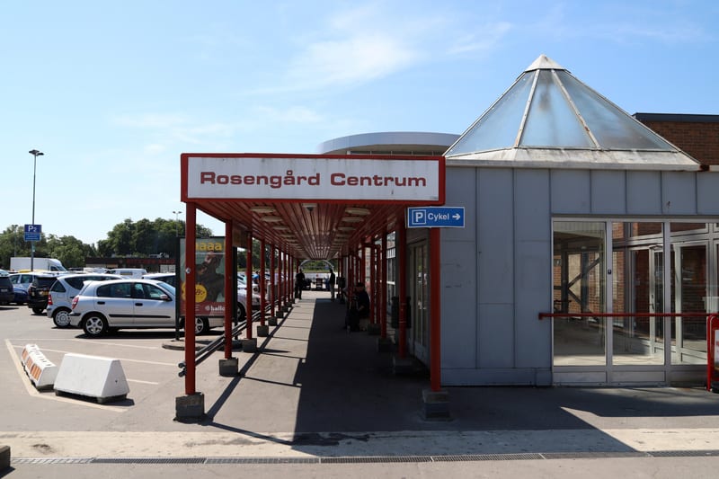 En utomhusbild som visar en parkeringsplats med flera parkerade bilar och en del av en byggnad med en entré täckt av ett rött tak. En skylt med texten "Rosengård Centrum" och en annan skylt som pekar mot "Cykel" är synliga. Mellan byggnaden och parke