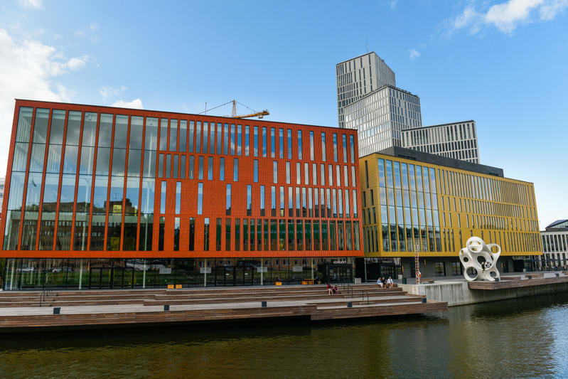 En bild som visar en orangefärgad, en gul och flera gråskaliga byggnader med stora glaspartier under en blå himmel med vita moln. I förgrunden syns en bred träbrygga längs en vattenyta. En gul byggkran syns i bakgrunden mellan två byggnader. En vit s