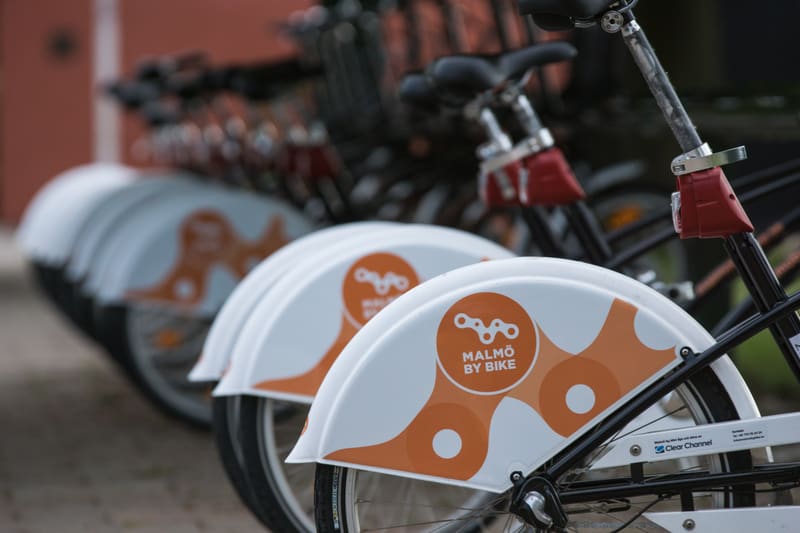 En bild som visar en rad parkerade cyklar från tjänsten "Malmö by Bike" med synliga stänkskydd som har orange logotyper och text på en vit bakgrund. Den främre cykeln är i fokus, medan de bakre cyklarna är oskarpa.