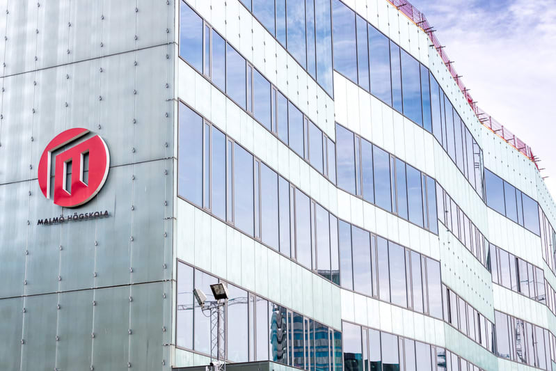 En modern byggnad med en fasad av glas och ljusa paneler. På byggnadens framsida syns logotypen för "MALMÖ HÖGSKOLA", röd med vita bokstäver. Fönstren reflekterar himmel och en byggkran. Byggnaden har en oregelbunden form upptill.