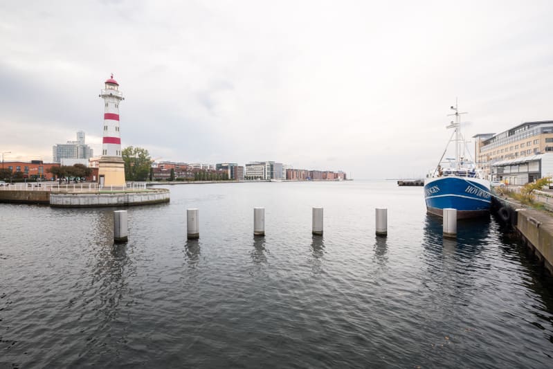 Bilden visar ett maritimt landskap med en röd- och vitrandig fyr på en cirkulär plattform i förgrunden till vänster. Till höger ligger en blå fiskebåt förtöjd vid en kaj. Bakom fyren och längsmed vattnet syns moderna byggnader i olika färger. Vattnet