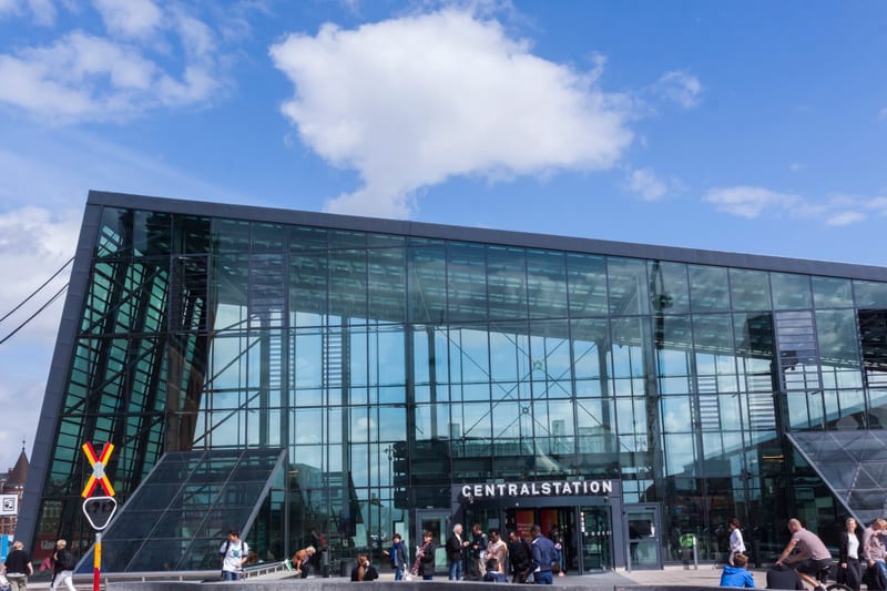 En modern byggnad med glasfasad och texten "CENTRALSTATION" ovanför entrén. Framför byggnaden syns flera människor och ett järnvägskorsningsmärke. Himlen är blå med några vita moln.