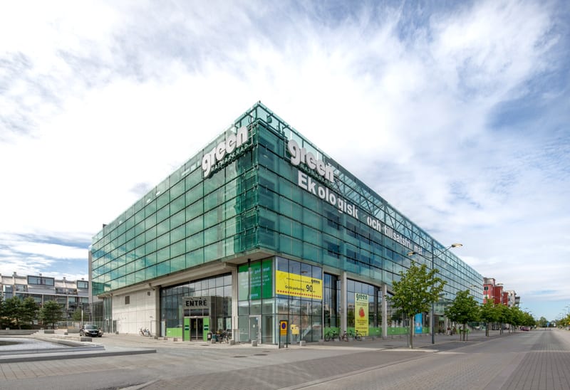 En modern byggnad med en fasad av glas och betong under en delvis molnig himmel. Byggnaden har skyltar med texten "green" och "Ekologisk och tillsatsfri mat". Framför byggnaden syns en trottoar, en gata och några träd. Ett par människor syns vid entr