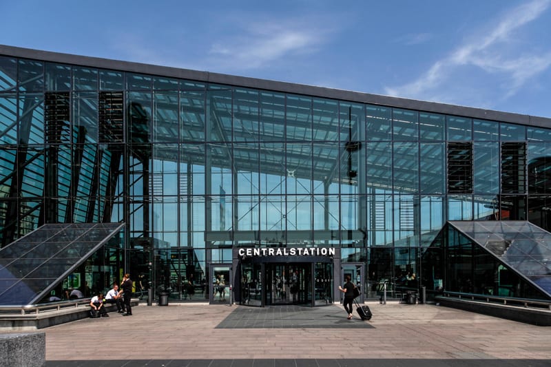 En modern byggnad av glas och metall med texten "CENTRALSTATION" ovanför entrén syns under en blå himmel. Framför byggnaden står och sitter flera personer, och en person med resväska är på väg mot entrén.