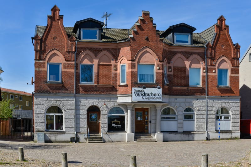 En fasadbild av en större tegelbyggnad med grå stengrund och en skylt för "Torget Vandrarhem & Logihotell". Byggnaden har många fönster i varierande storlekar, spröjsade takkupor på taket, samt flera dörrar och trappor. Förgrunden består av en stenla