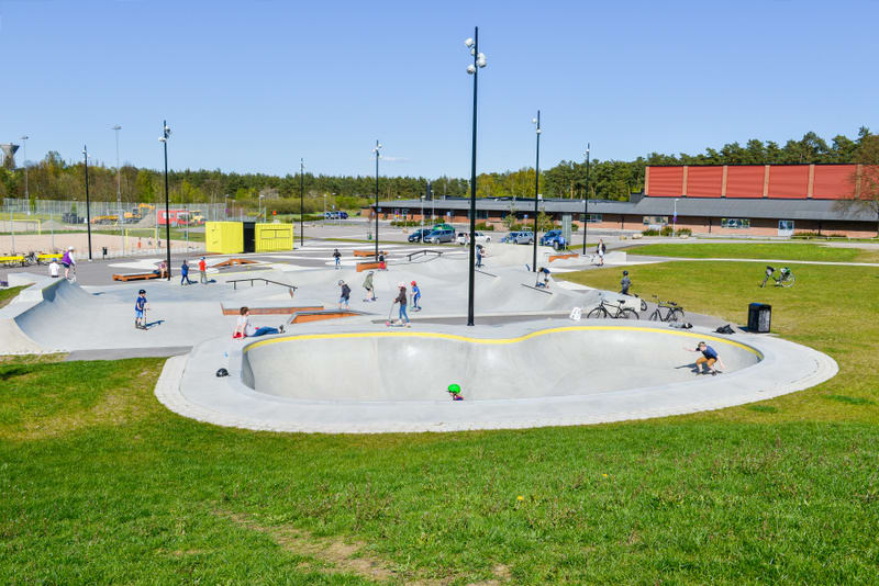 En översiktsbild av en skateboardpark med flera ramper och banor. Ett flertal personer syns i parken, varav många åker skateboard eller sparkcykel. Gröna gräsmattor omringar parken. I bakgrunden syns bilar på en parkeringsplats, byggnader och träd.