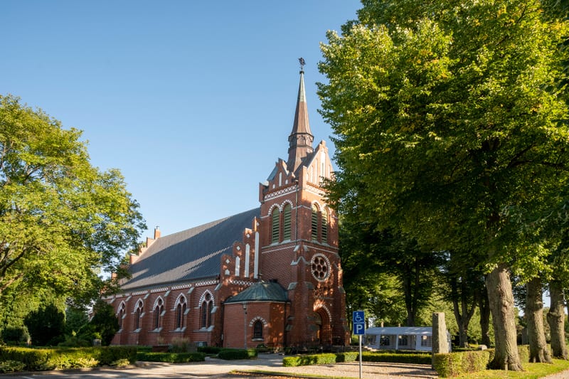En röd tegelkyrka med spetsigt torn och detaljerade fönster står omgiven av lummiga gröna träd under en klarblå himmel. Ett parkeringsskylt för rörelsehindrade syns i förgrunden till höger, intill ett vitt tält.