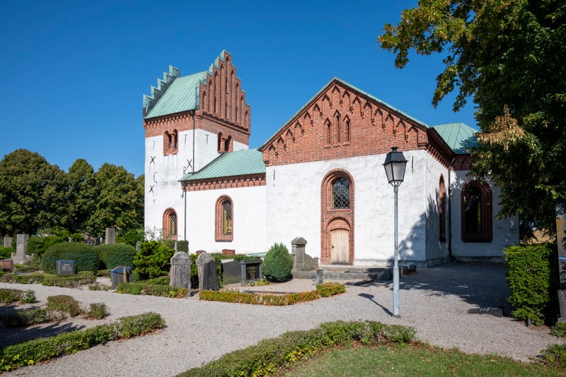 En kyrka med vitmålade väggar, röda tegeldetaljer och grönt tak. Framför kyrkan syns en kyrkogård med gravstenar, grusgångar och buskar. Himlen är klarblå och solen bidrar till skarpa skuggor.