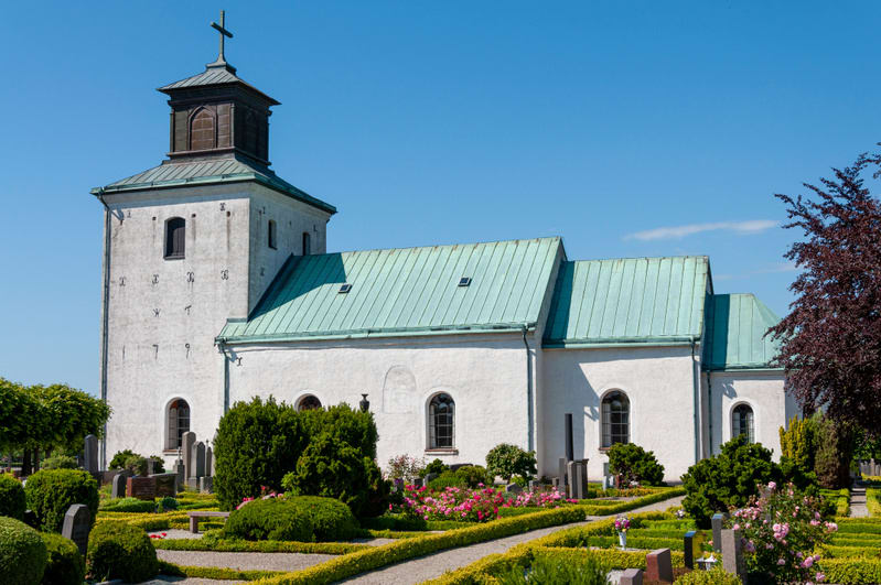 En vit kyrka med grönt tak under en klarblå himmel. Runt kyrkan finns en grönskande kyrkogård med buskar, blommor och gravstenar. Solen lyser på kyrkan och kastar skuggor, vilket indikerar att det är dagtid.