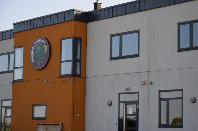 En modern byggnad med en fasad i två färger: en orange sektion till vänster och en ljusare, nästan vit, sektion till höger. På den orangea sektionen finns en stor rund skylt med texten "BARSEBÄCKS MONTESSORI" och en stiliserad bild av ett träd. Flera
