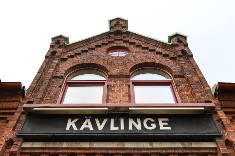 En fasad på en röd tegelbyggnad med två välvda fönster och ett runt fönster ovanför dem. Under de större fönsterna finns en svart skylt med vit text "KÄVLINGE". Taket har en grön kant och himlen i bakgrunden är ljus.