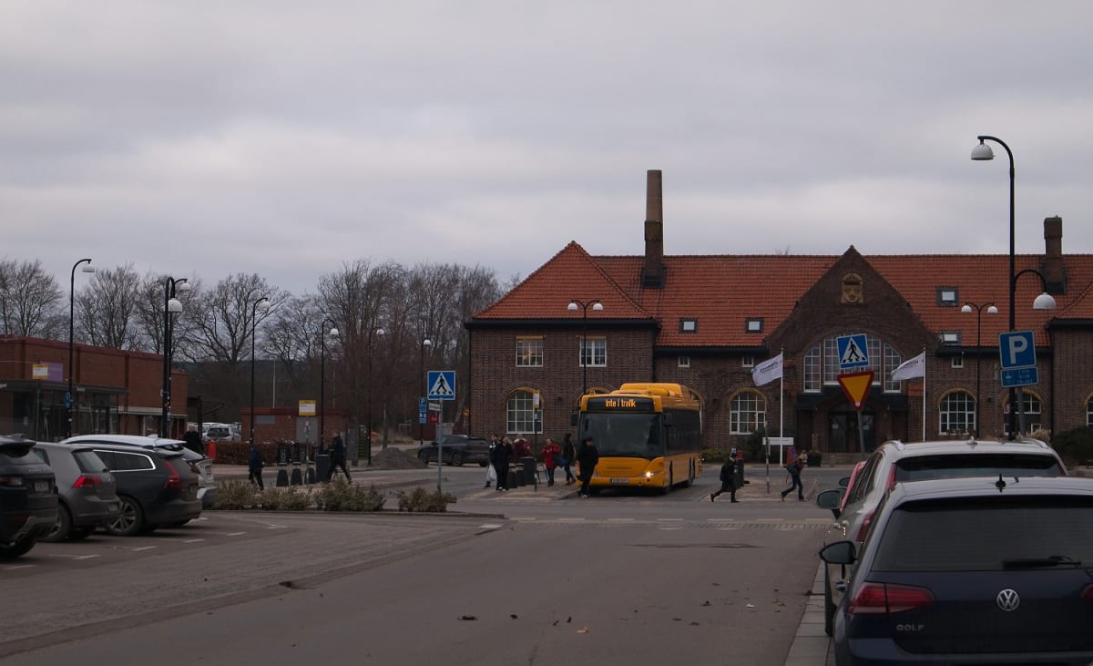 En gul buss står utanför en mörk tegelbyggnad med rött tegeltak. Flera personer rör sig på trottoarer och över ett övergångsställe. Personbilar står parkerade längs vägkanten. Himlen är grå och molnig.