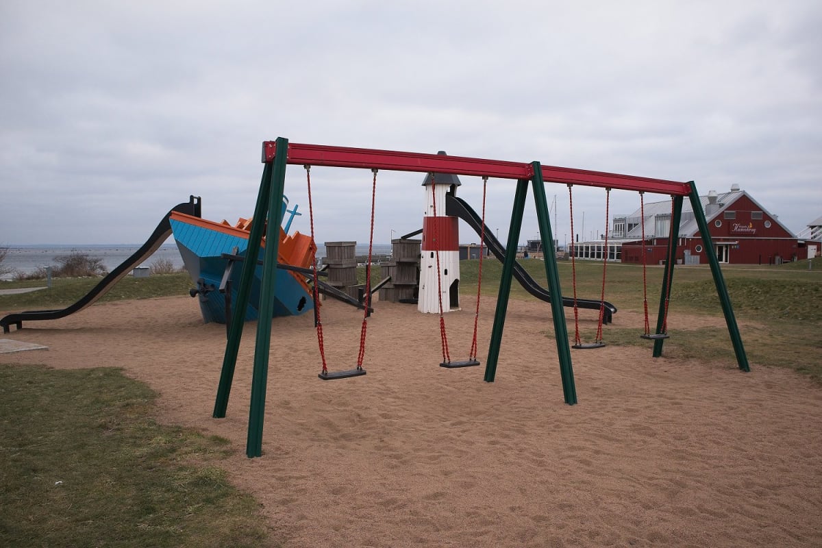 En lekplats med sandunderlag och gräs i förgrunden, omgiven av byggnader och vatten i bakgrunden under en gråmulen himmel. I mitten står en gunga med sex sitsar, och till vänster syns en båtformad lekställning med svart rutschkana bredvid en fyrforma