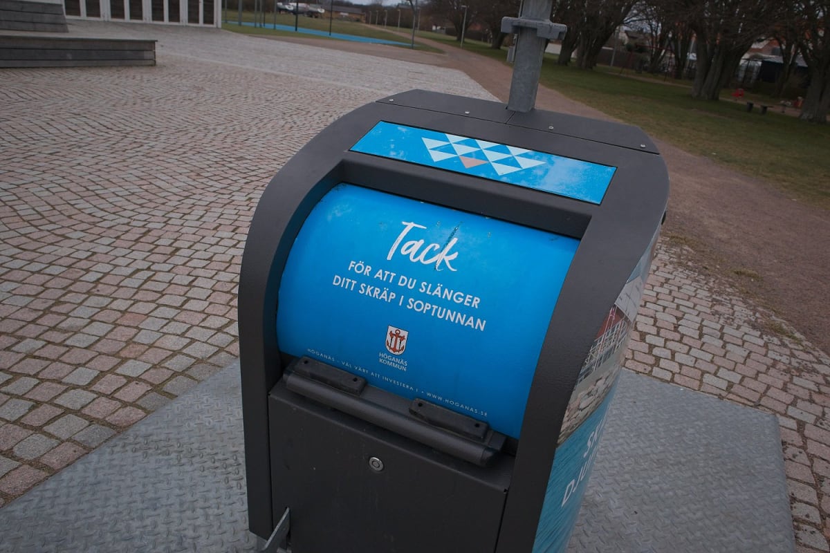 En svart och blå soptunna står på marken. Texten "Tack FÖR ATT DU SLÄNGER DITT SKRÄP I SOPTUNNAN" är synlig på den blå ytan. En logotyp för Höganäs kommun är också avbildad. Bakgrunden består av en stenlagd yta, gräs och träd.