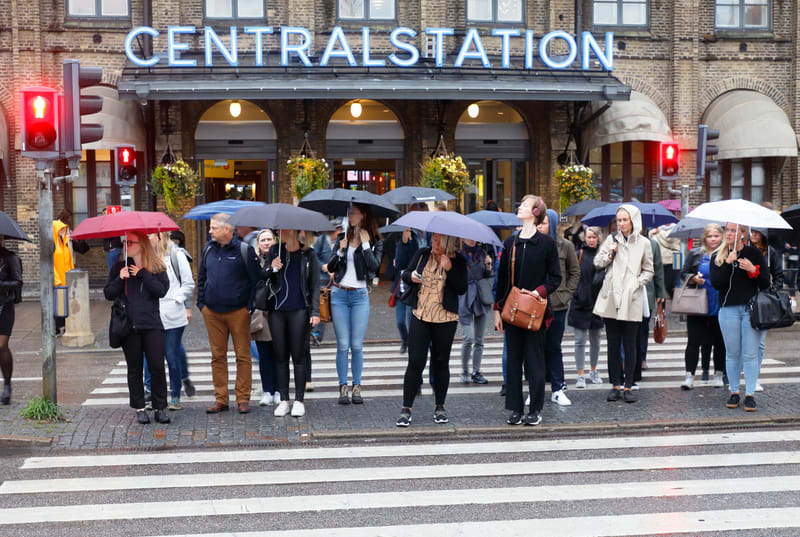 En grupp människor står på en övergångsställe nära en byggnad med skylten "CENTRALSTATION". Många håller upp paraplyer, vissa röda, svarta, vita, eller blåa. Två trafikljus med röda lampor syns. Byggnaden bakom människorna är i tegel med valvformade