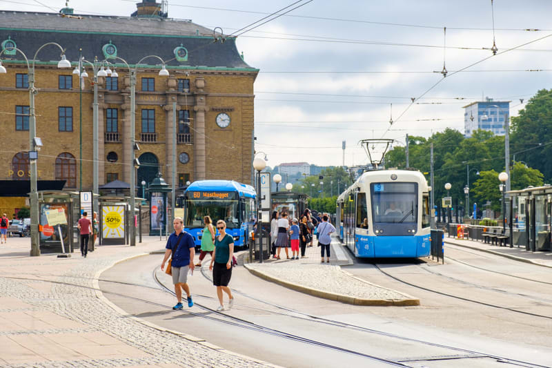 En gatuscen från en stad i dagsljus, där en blå buss och en blåvit spårvagn står vid hållplatser. Flera människor syns gående och väntande. I bakgrunden finns en gul tegelbyggnad med texten "POST" och en klocka. Grönska och en modern byggnad syns i f
