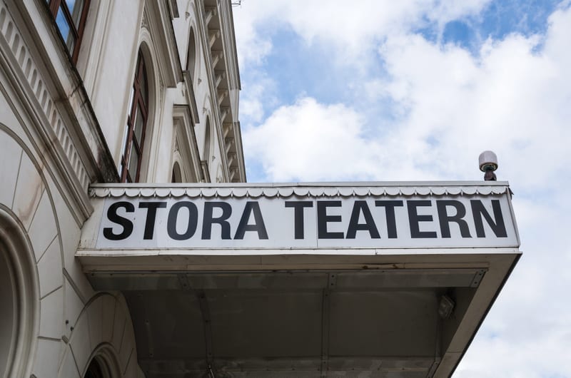 En bild av delen av en byggnadsfasad med en skylt det står "STORA TEATERN" på. Skylten är vit med svarta bokstäver och har en lampa på ena sidan. Bakom skylten syns den vita byggnaden med fönster och en blå himmel med moln.