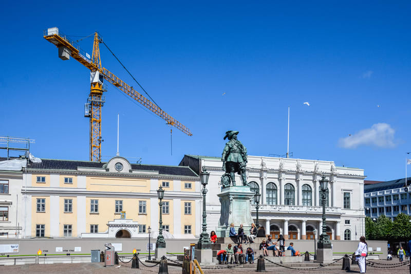 En stor, horisontell bild visar ett torg med en staty i förgrunden, omgiven av människor som sitter på trappsteg. I bakgrunden syns flera byggnader. Ett gult byggnadskran med blå himmel syns ovanför byggnaderna. En svart kedja löper längs basen av st