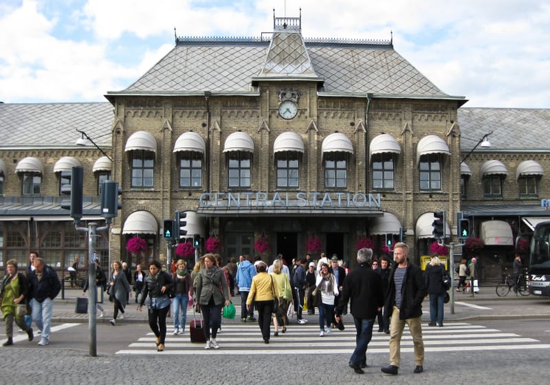 En stor byggnad med gul tegel och grått tak, märkt "CENTRALSTATION", syns i bakgrunden. Framför byggnaden finns ett övergångsställe med många människor som korsar gatan. Trafikljus med grönt sken är synliga.